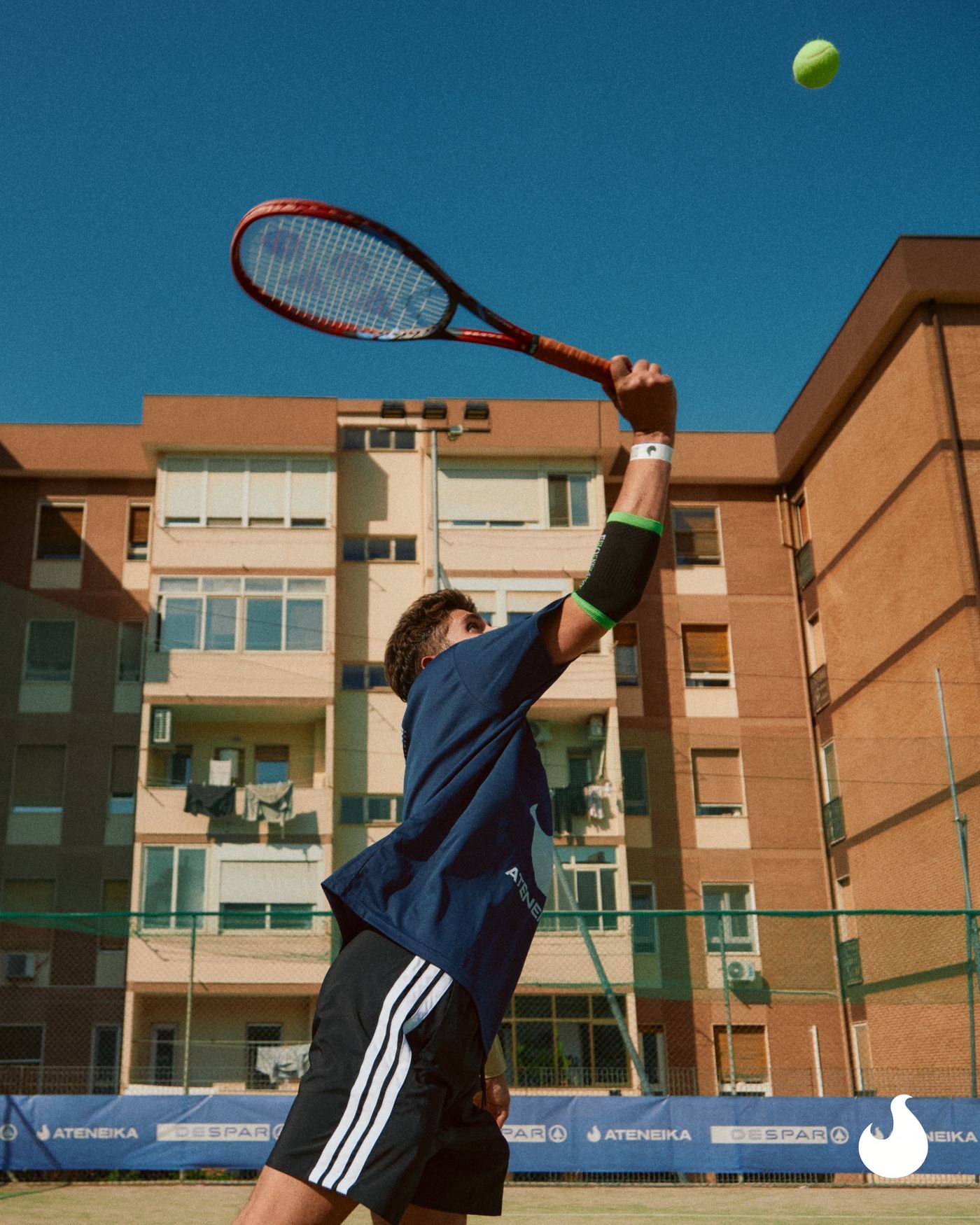 Un ragazzo al servizio sul campo da tennis, con i palazzi sullo sfondo.