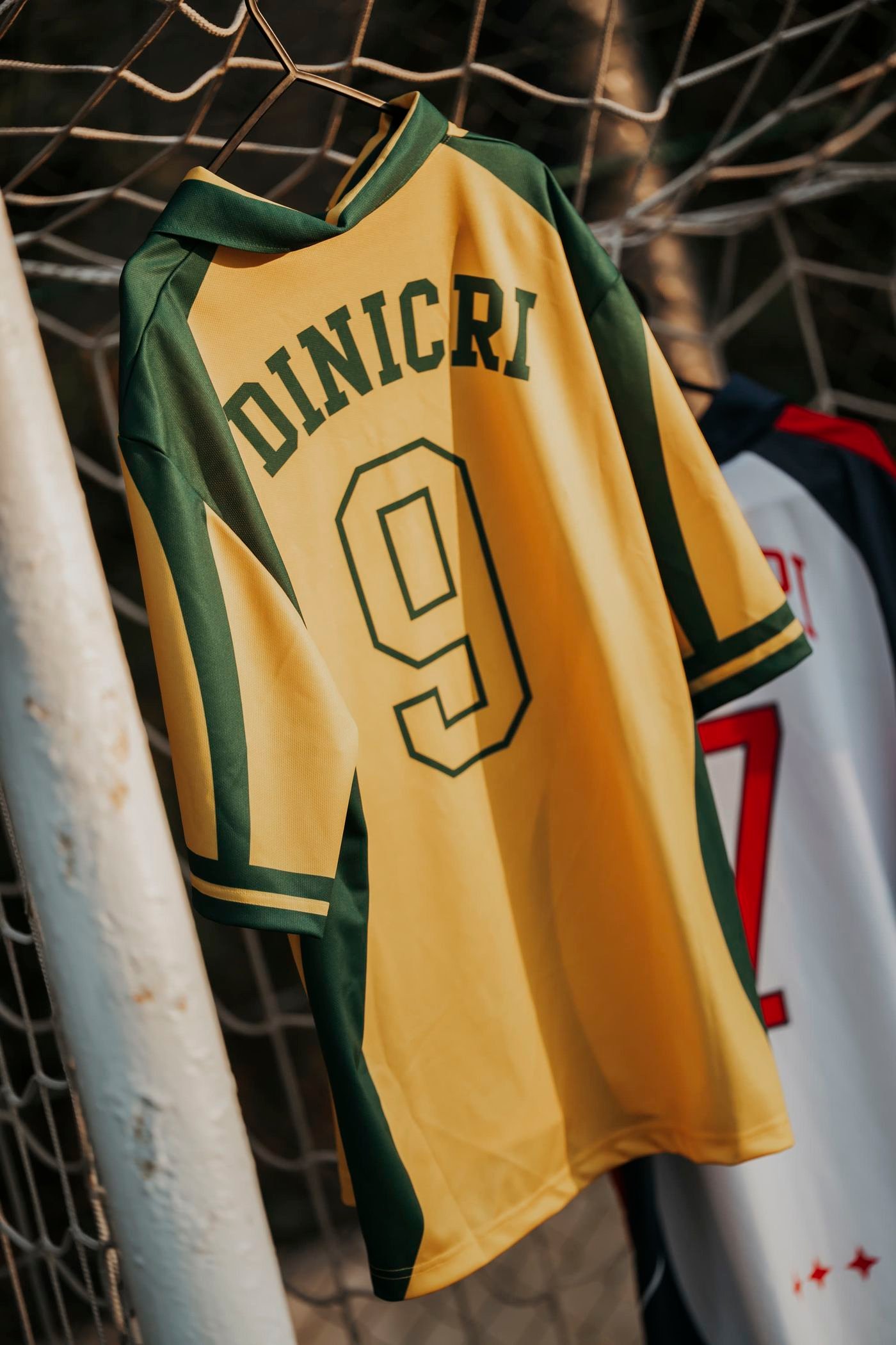 Una maglia gialla e verde della collezione Dinicri, appesa a una gruccia sulla rete di una porta da calcio.