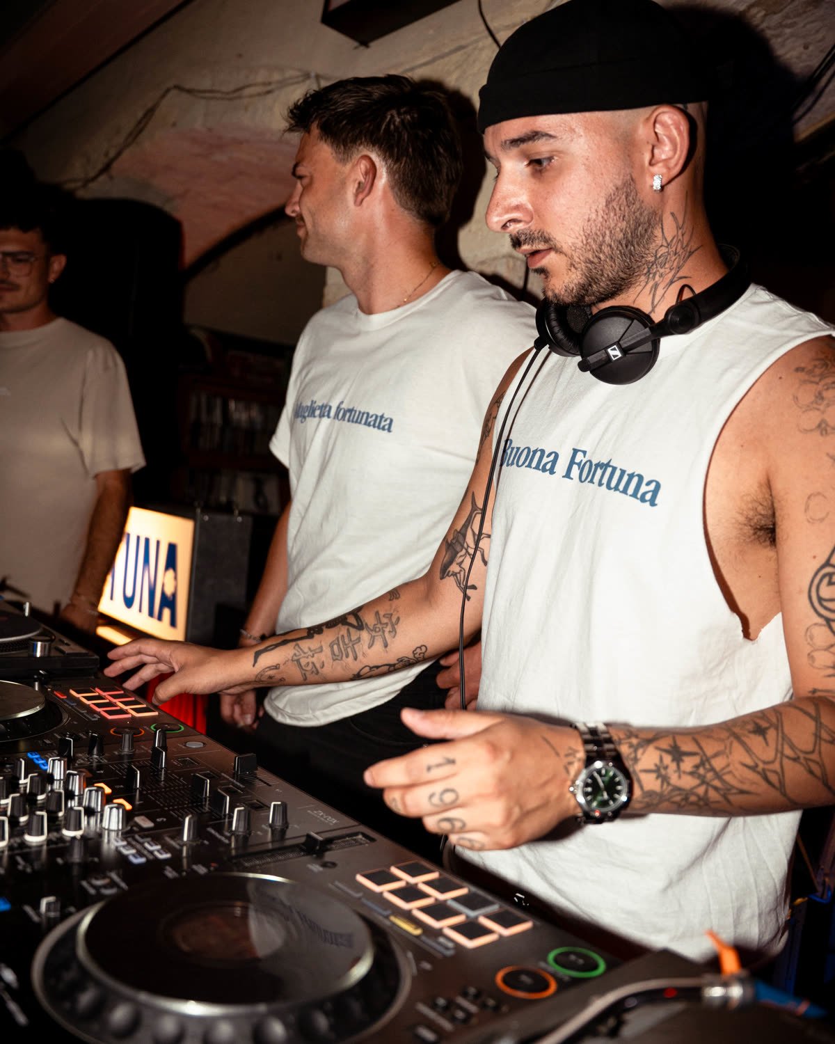 Due dj alla consolle con la canotta bianca e la scritta Buona fortuna in blu sul petto.
